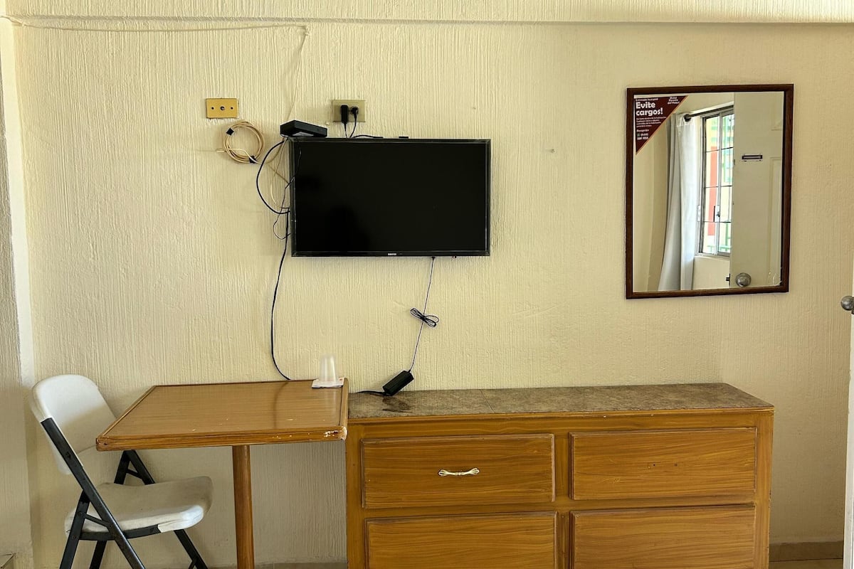 Une pièce comprenant un bureau en bois, une chaise, un téléviseur fixé au mur, un miroir et une courtepointe posée sur le sol.