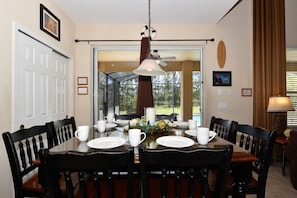 Une salle à manger avec une grande table dressée pour six personnes, entourée de chaises en bois foncé. Un ventilateur de plafond se trouve au-dessus de la table, et une fenêtre avec des rideaux est située à gauche.