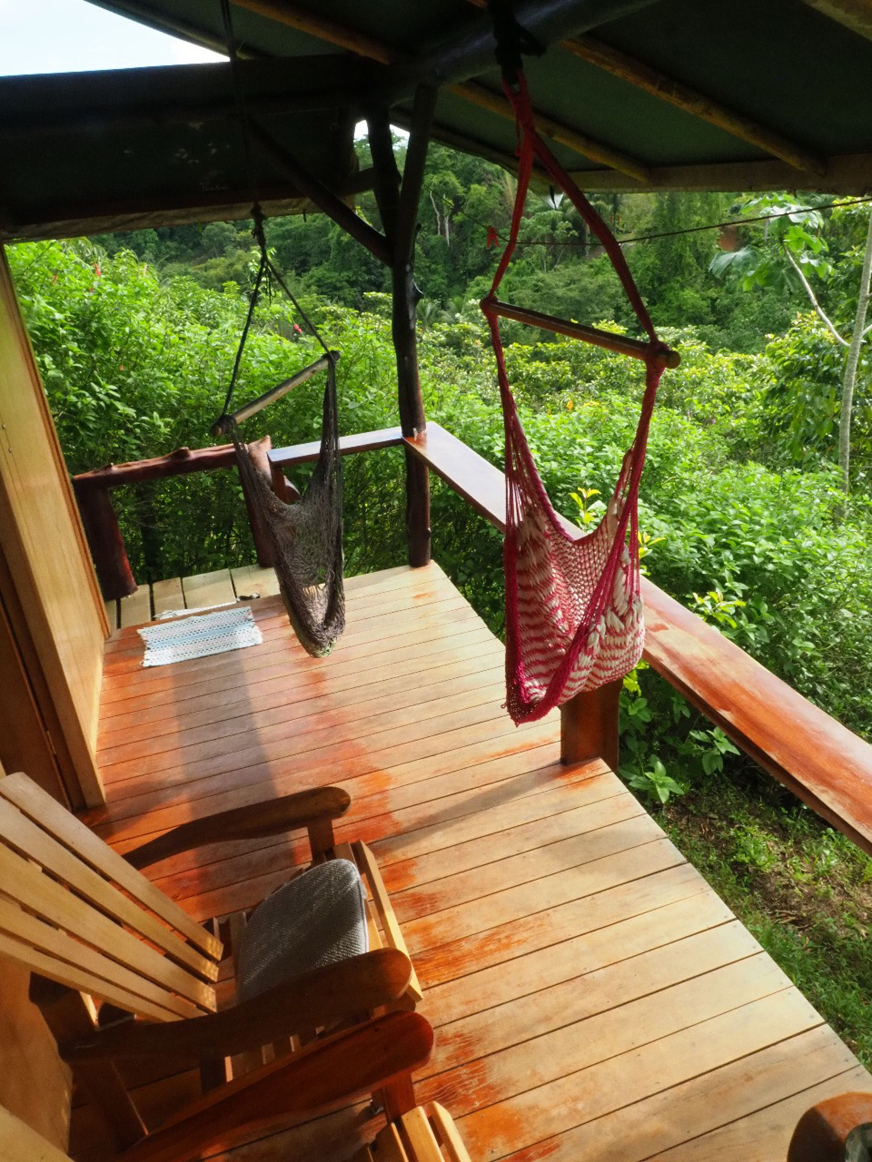Una terraza de madera con dos columpios y una silla, con vista a un bosque verde y frondoso.