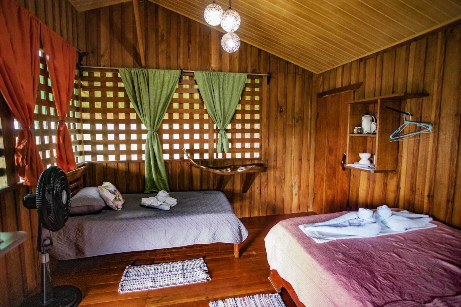 A wooden cabin room with two beds, a fan, a woven mat, and a shelf with toiletries.