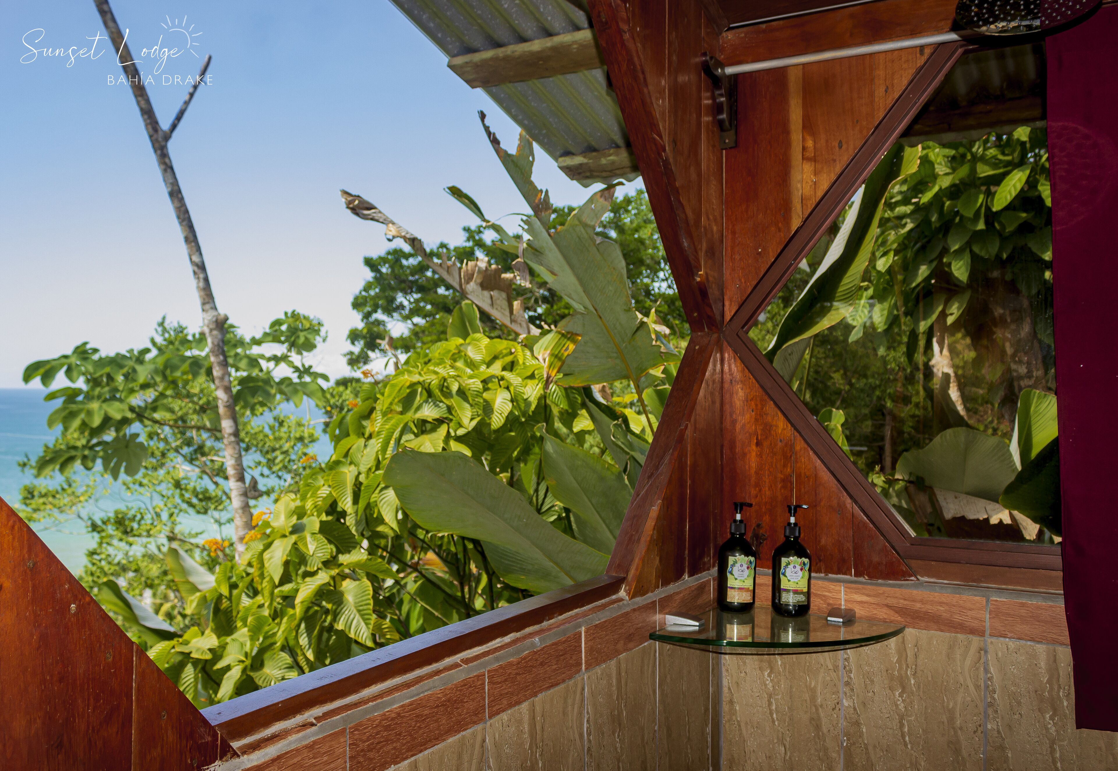 A corner of a room with a wooden ceiling, a glass table with two bottles, and a view of the ocean and tropical foliage.