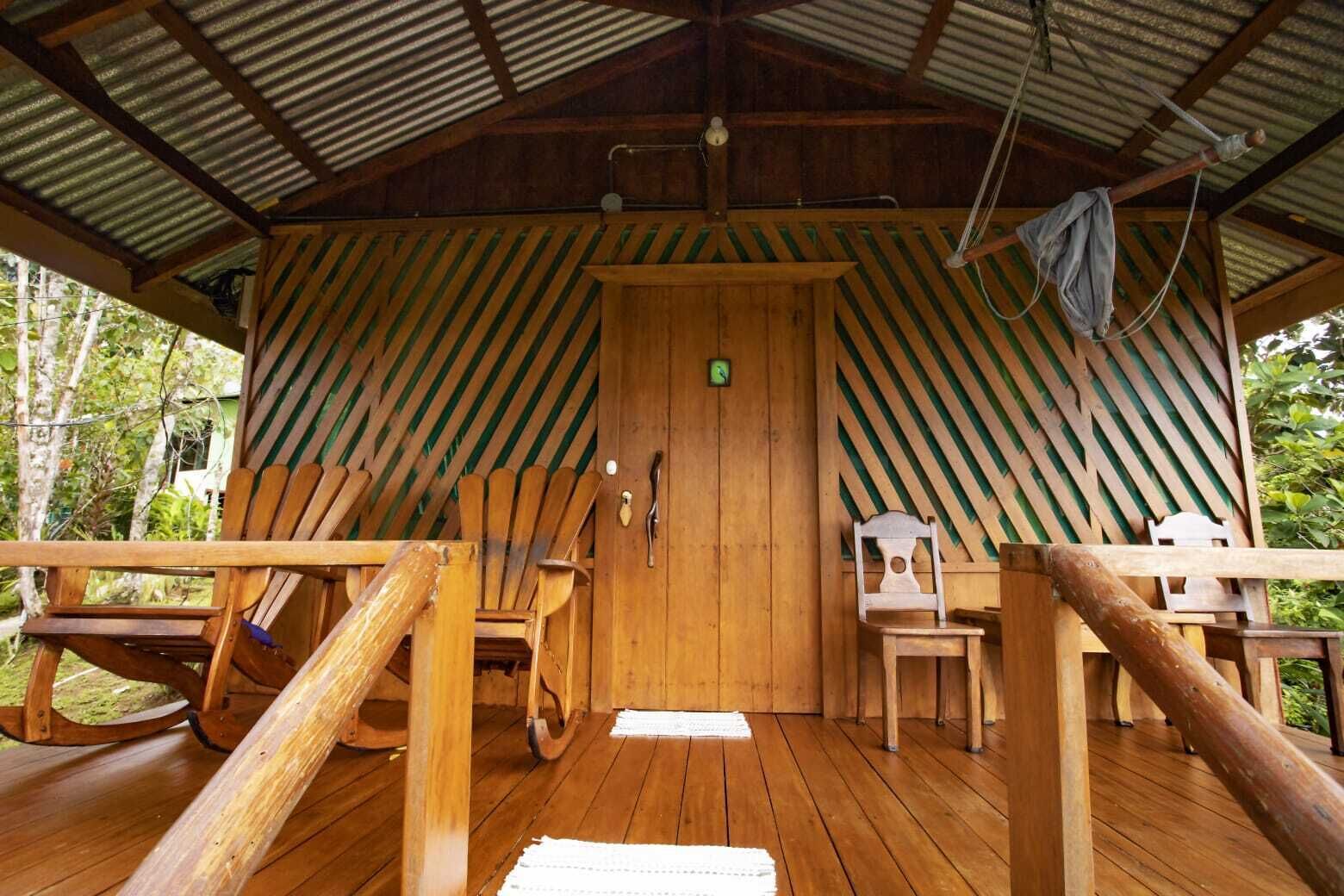 Una cabaña de madera con techo de metal corrugado, puerta de madera y un porche con muebles de madera, incluyendo mecedoras y una mesa.