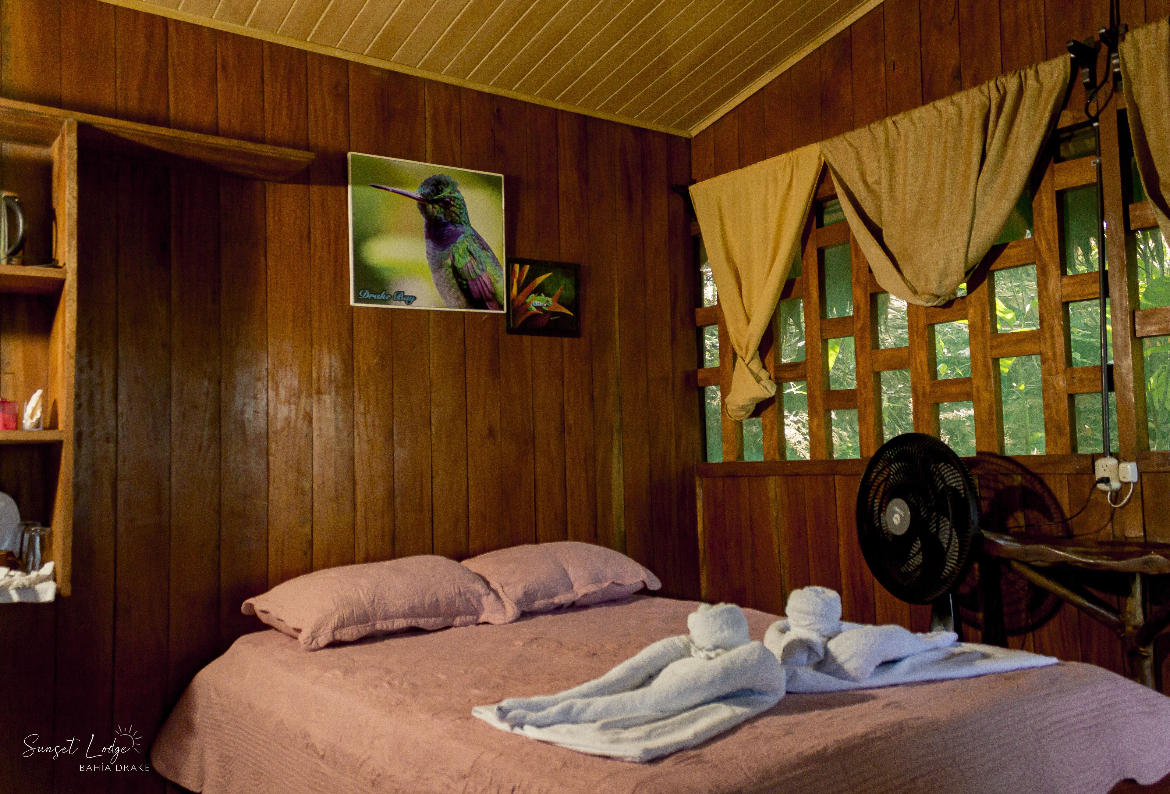 A wooden room with a bed, pillows, a fan, and a window with curtains.