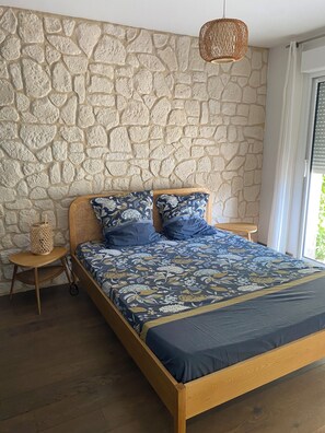 Une chambre avec un mur en pierre, un lit en bois, une lampe en osier et une petite table en bois.