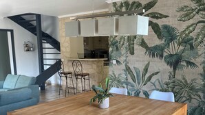 A dining area with a wooden table, white chairs, a bench, a potted plant, a kitchen in the background, and a staircase.