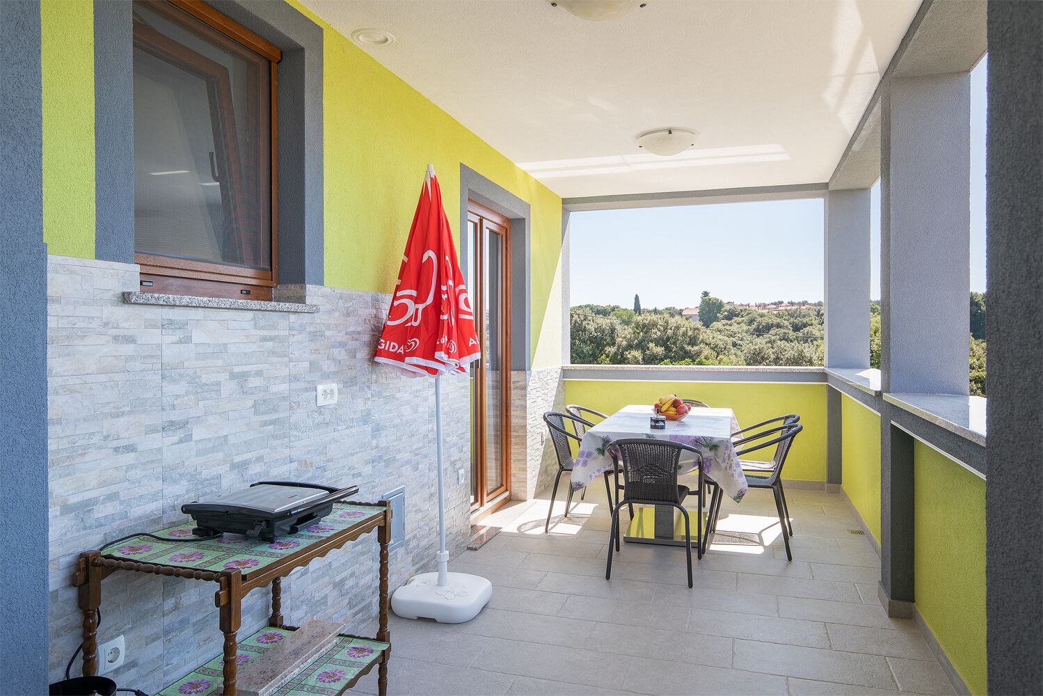 A balcony with a table and chairs, a red umbrella, and a view of trees and hills.