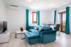 Ein modernes Wohnzimmer mit einer Eckcouch, einem Flachbildfernseher, einem Essbereich mit Tisch und Stühlen sowie einem Balkon mit Vorhängen.