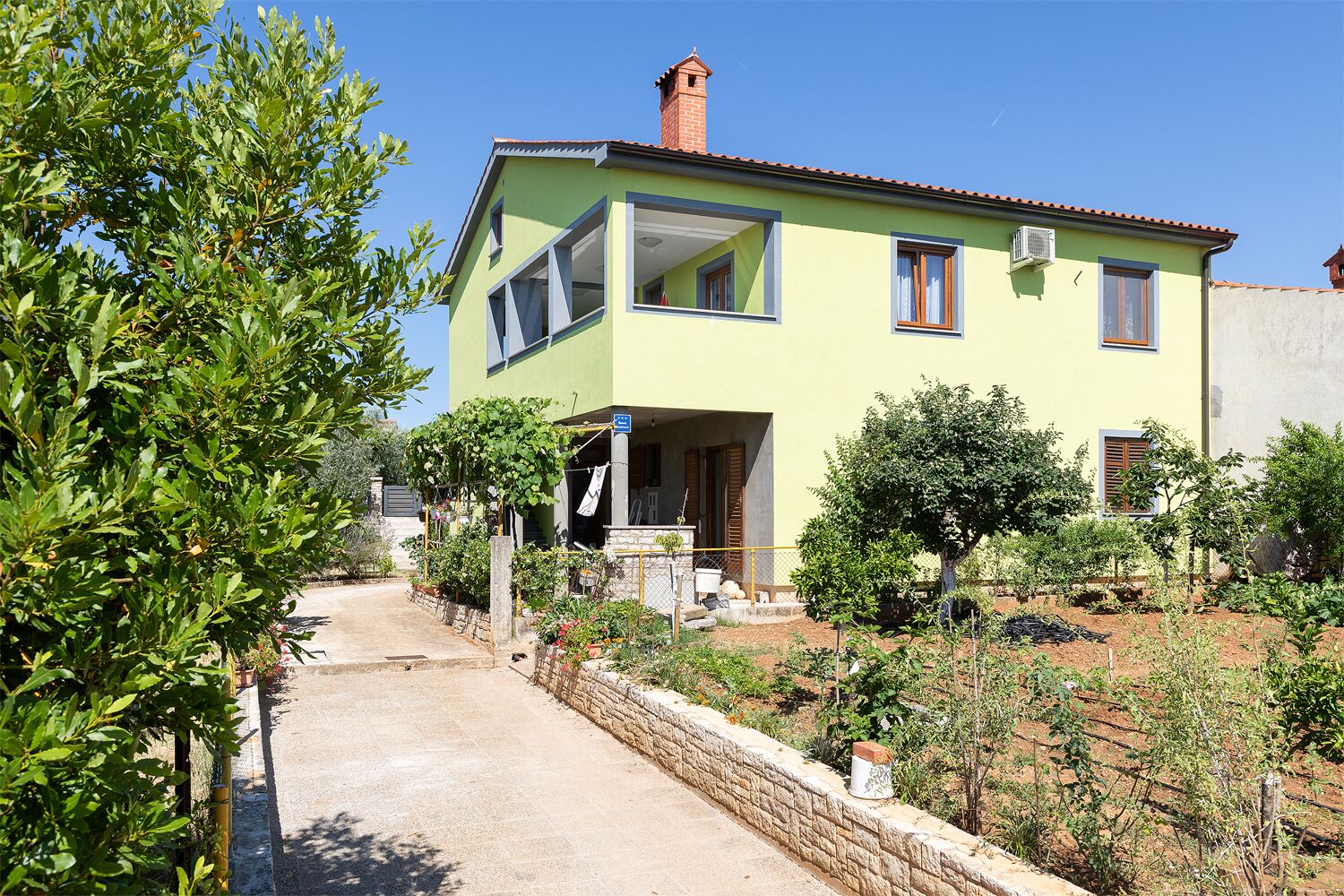 Une maison à deux étages avec une façade verte, une cheminée bien visible et un mur de pierre au premier plan.