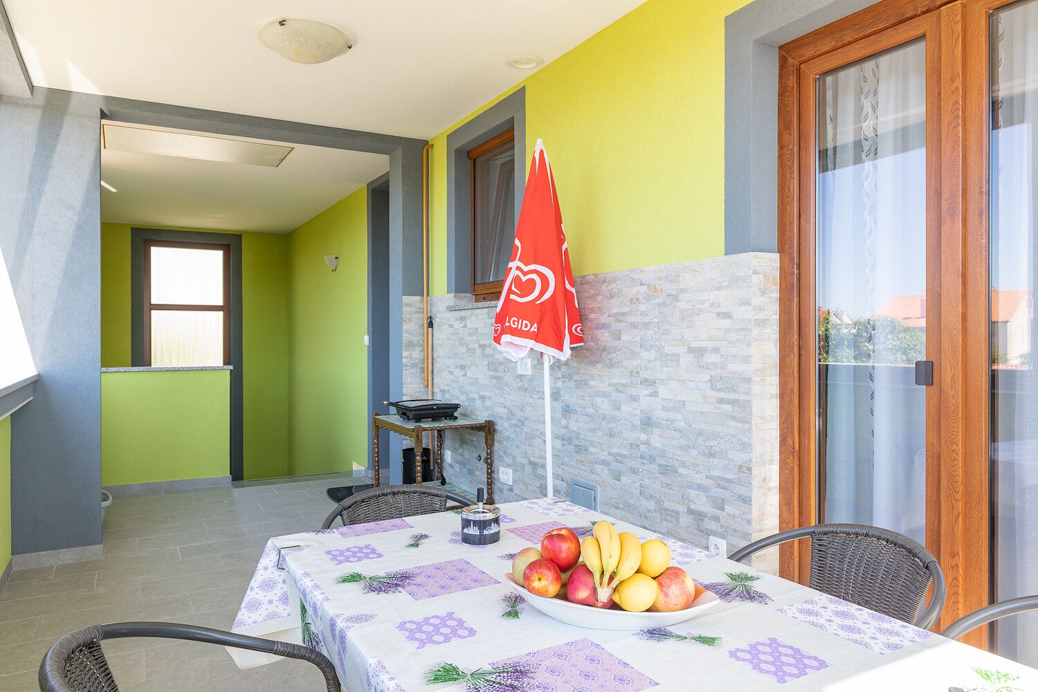 A dining area with a table set for two, a bowl of fruit, and a red umbrella with a logo.