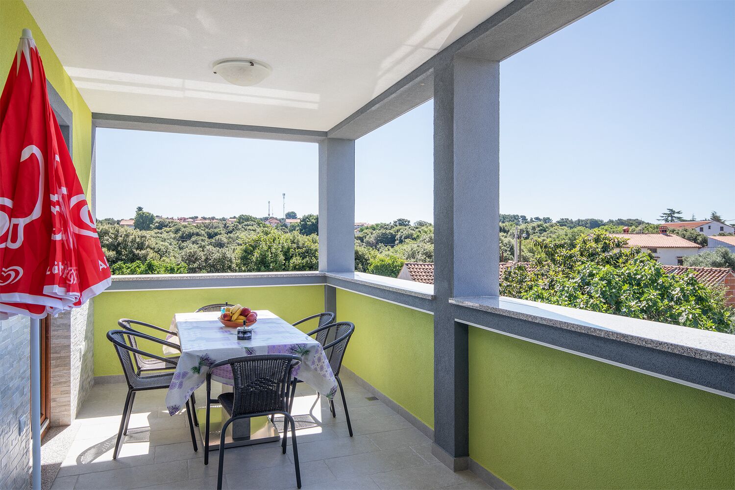 Un balcon avec une table et des chaises, un parasol rouge, et une vue sur la verdure et les bâtiments.