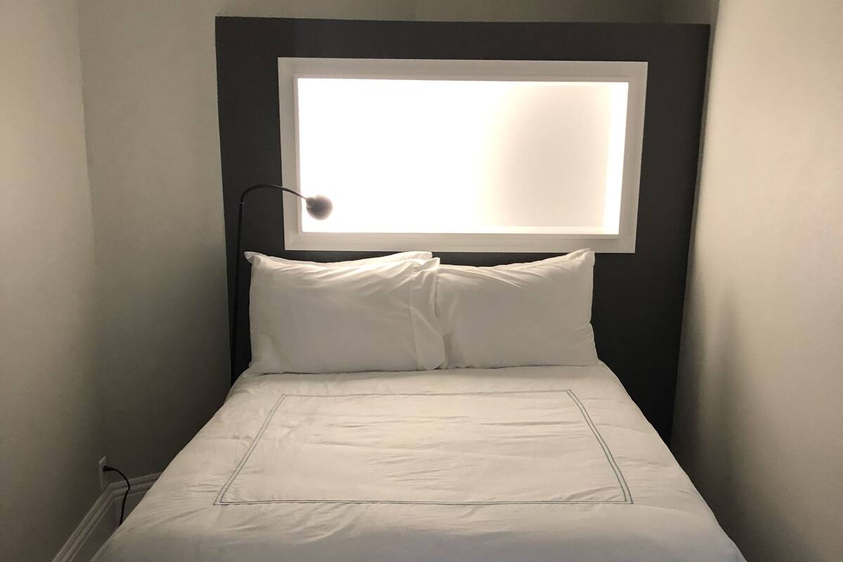 Una cama bien tendida con cabecera, una lámpara de noche y un enchufe en la pared.