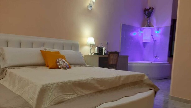 A bedroom with a large bed, a nightstand, a lamp, and a bathroom with a purple-lit shower. - Modina Guest House (Rome)