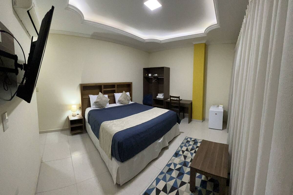 A hotel room with a bed, a desk, a chair, a small table, a wardrobe, a mirror, a television mounted on the wall, and a patterned rug.