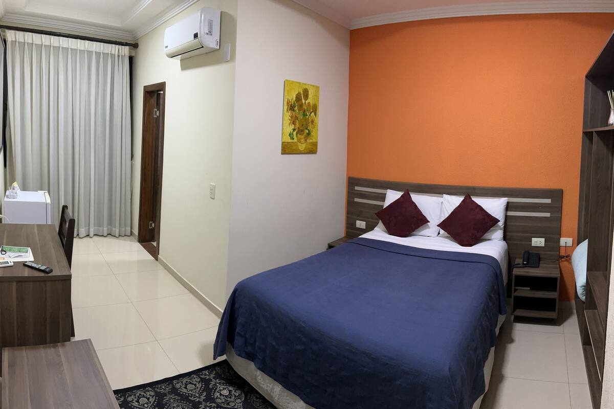 A hotel room with a bed, a desk, a chair, a TV, and a window with curtains.