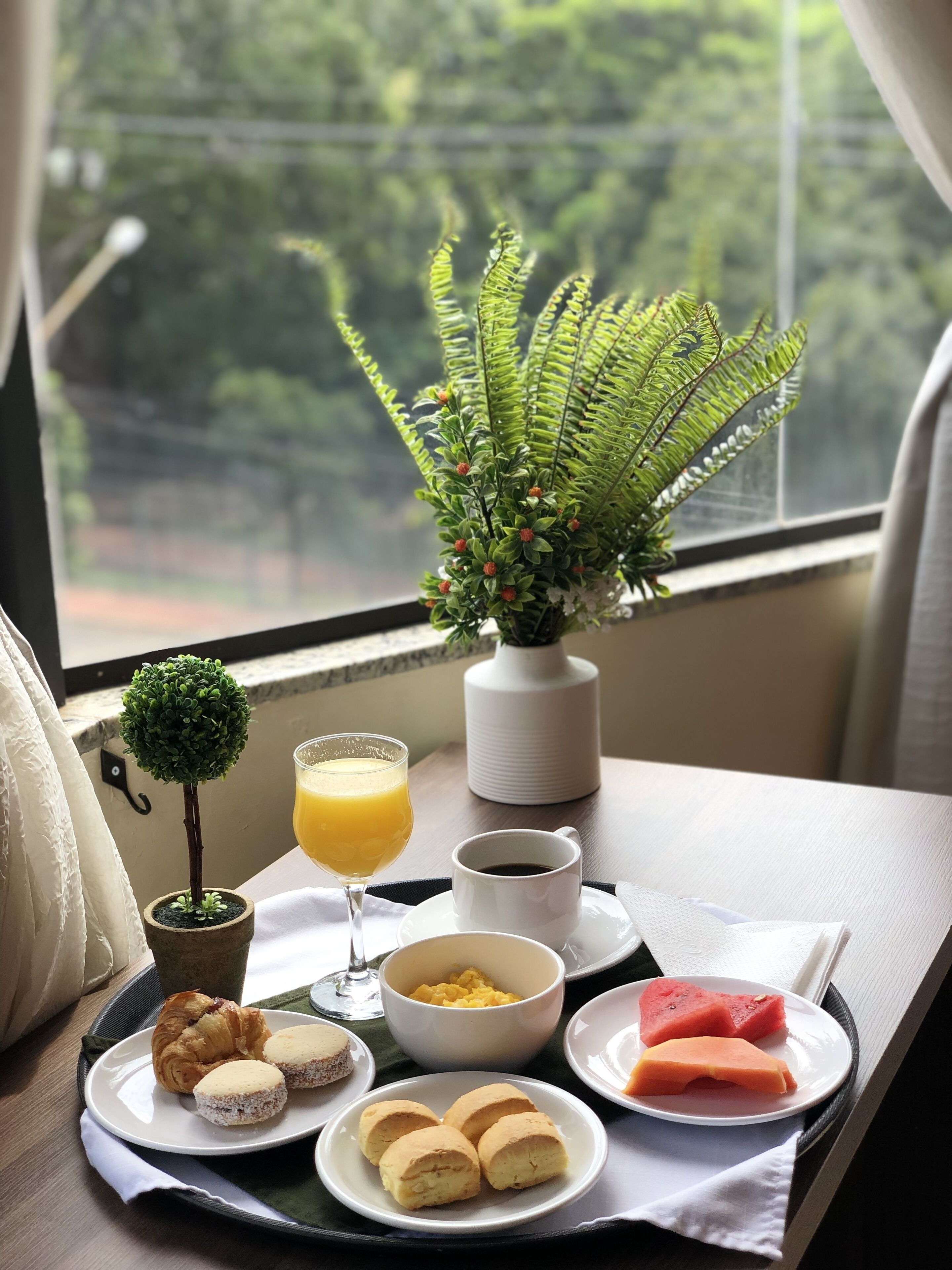 Bandeja de café da manhã com croissants, biscoitos, melancia e suco de laranja sobre uma mesa próxima a uma janela com uma planta e vista para o verde.