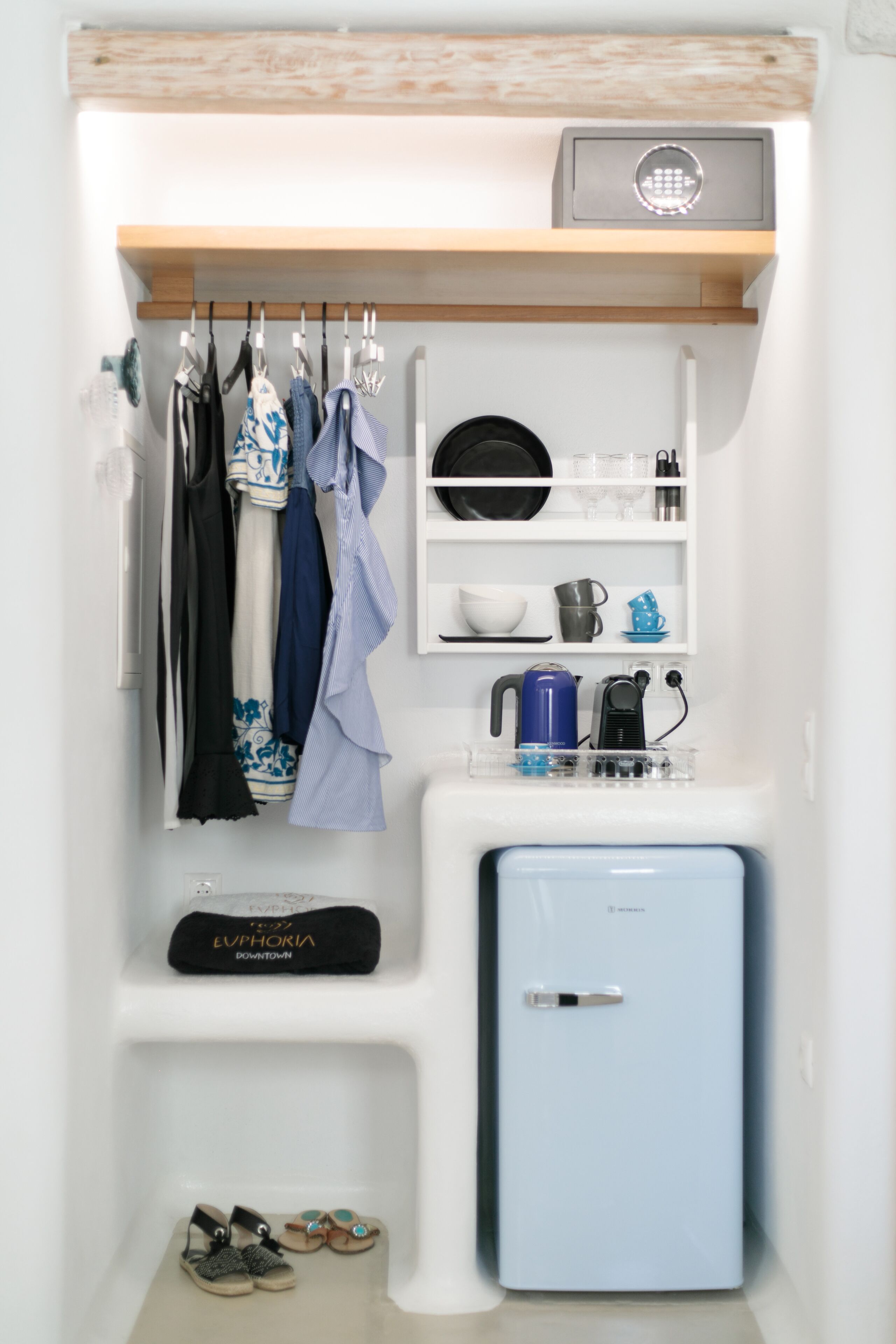 A compact kitchenette with a blue refrigerator, a coffee maker, and a kettle. Clothes are hung on a rack above, and a pair of sandals is visible on the floor.