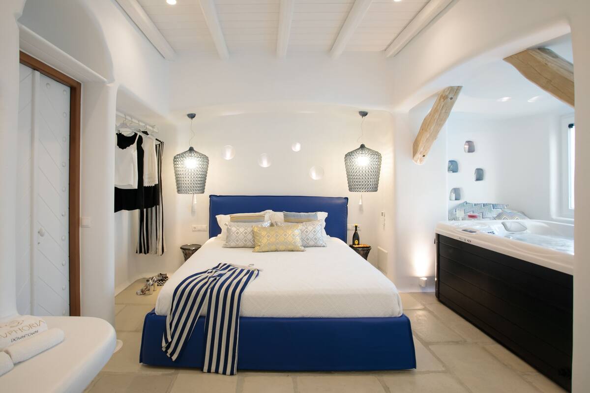 A modern bedroom with a large bed, bedside tables, a hanging light fixture, and a built-in bathtub.