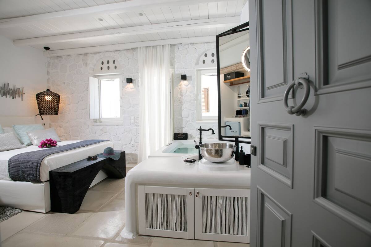 A modern bedroom with a stone wall, a bed with a bench, a white vanity with a mirror, and a bathroom visible through a window.