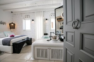 A modern bedroom with a stone wall, a bed with a bench, a white vanity with a mirror, and a bathroom visible through a window.