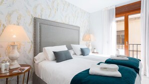Une chambre d’hôtel avec deux lits, une tête de lit en bois, du linge de lit blanc, des coussins décoratifs de couleur turquoise et une table de chevet en bois avec une lampe et un magazine.