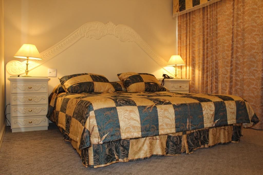Una cama bien hecha con una colcha a cuadros, flanqueada por dos mesitas de noche con lámparas, en una habitación con papel tapiz estampado y un medallón decorativo en el techo.