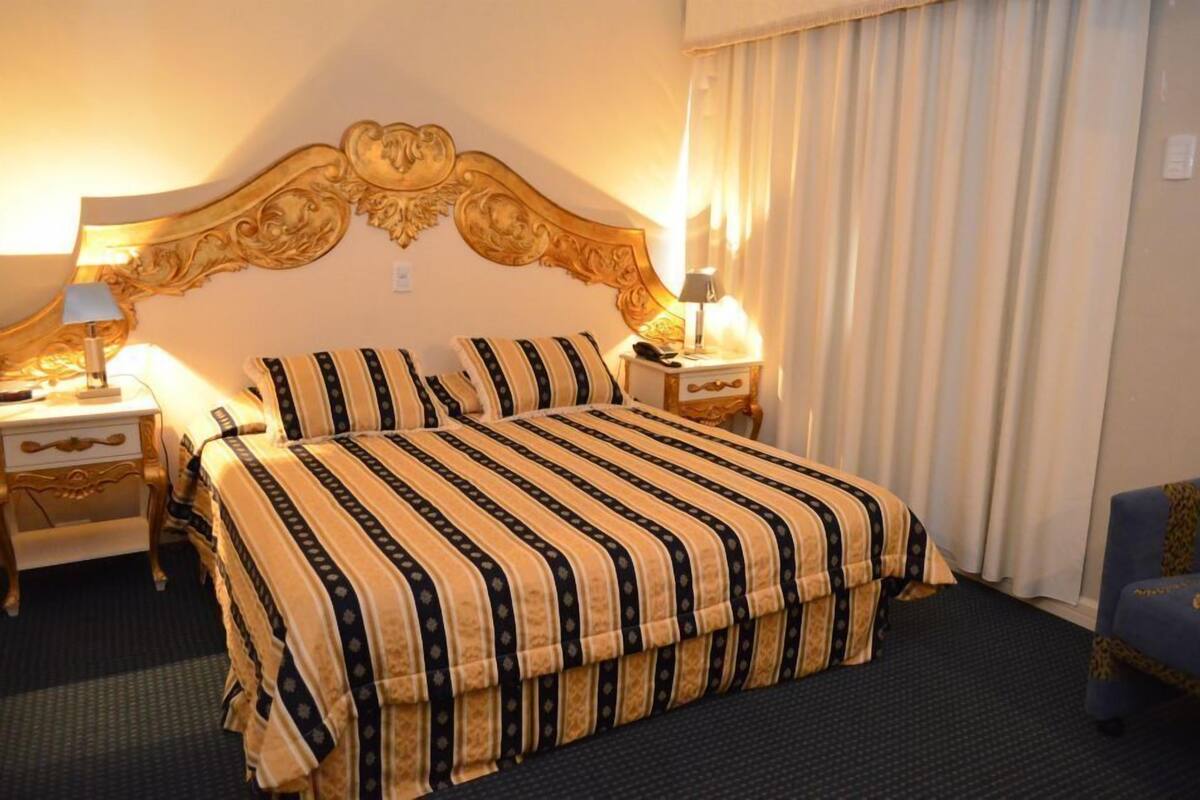 A hotel room with a large bed, two bedside tables with lamps, a blue armchair, and a decorative headboard.