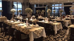 Une salle de banquet où les tables sont dressées pour un événement formel, agrémentée de grands vases remplis de fleurs blanches et de larges fenêtres offrant une vue sur le paysage urbain.