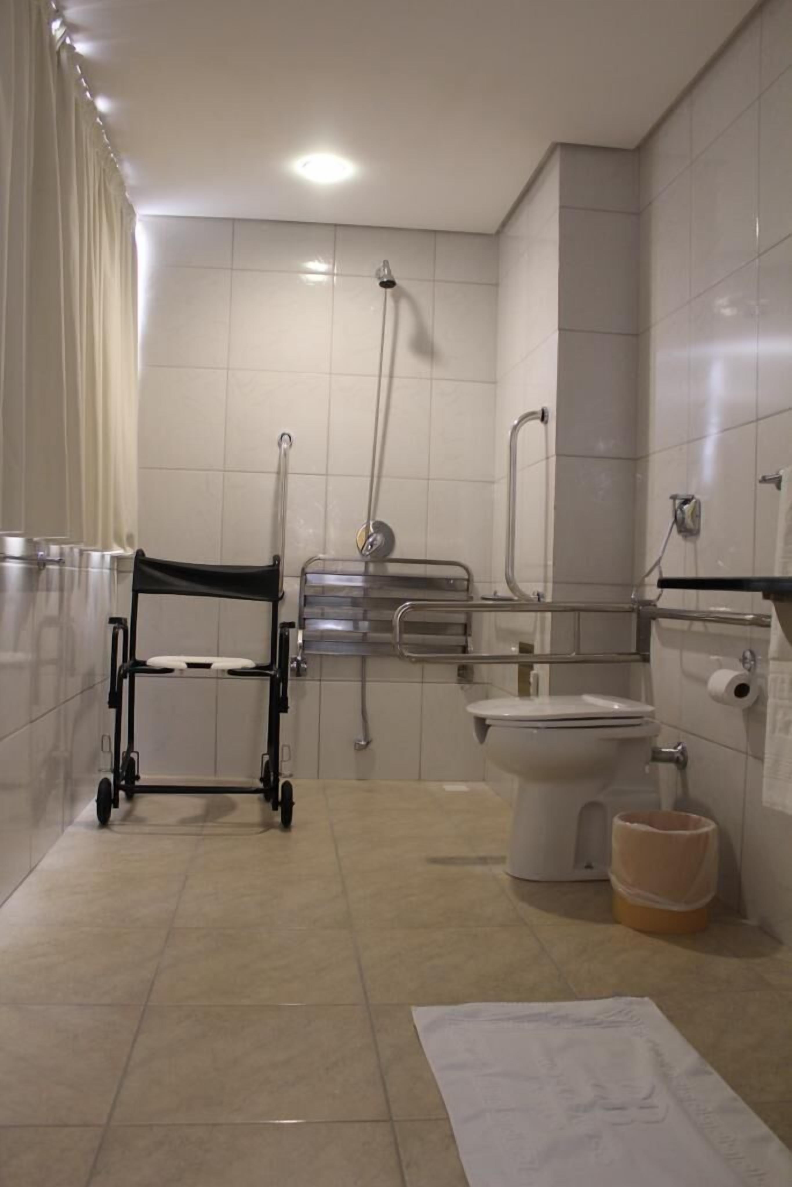 A bathroom with a wheelchair, a toilet, a shower, and a handrail.
