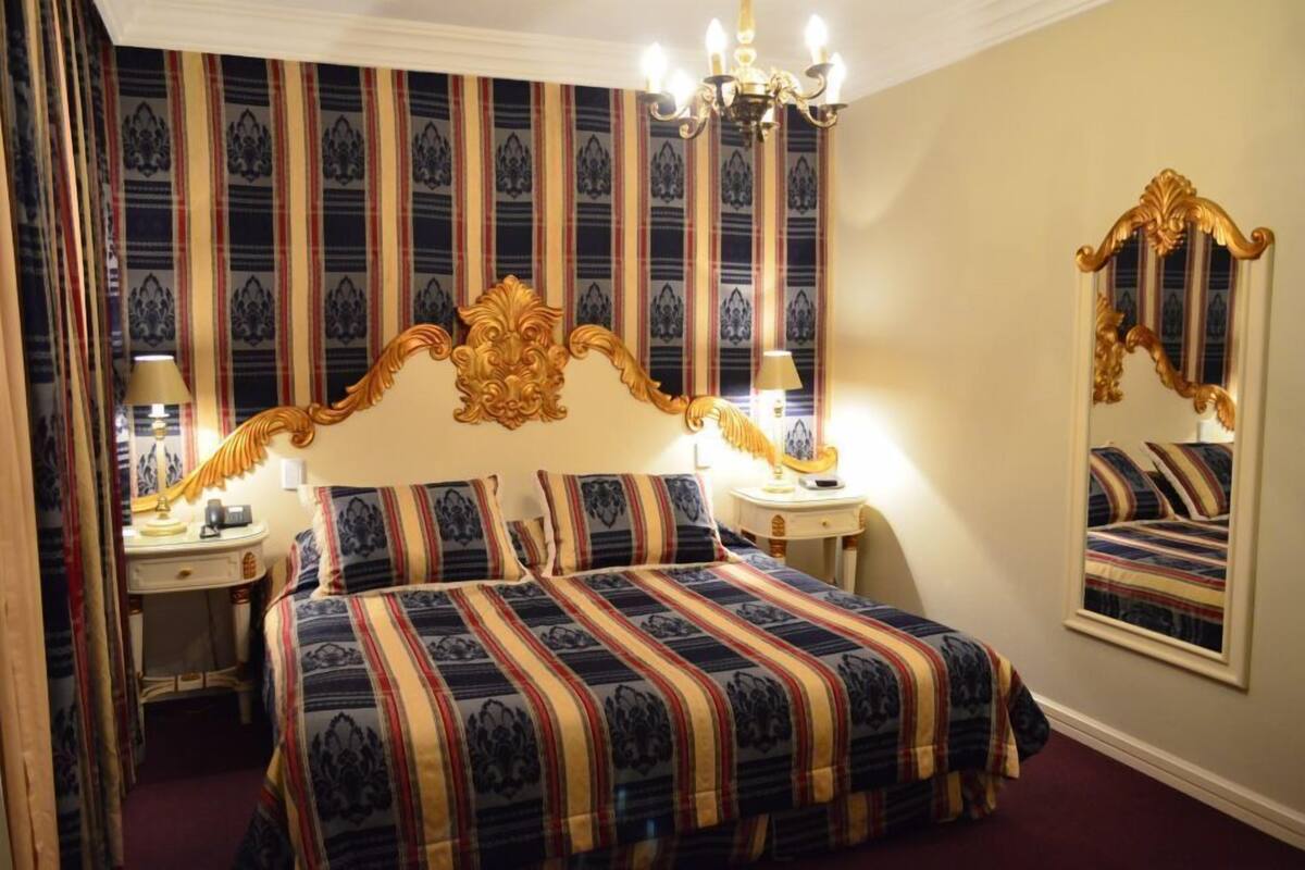 A hotel room with a large bed, a mirror, a chandelier, and striped wallpaper.