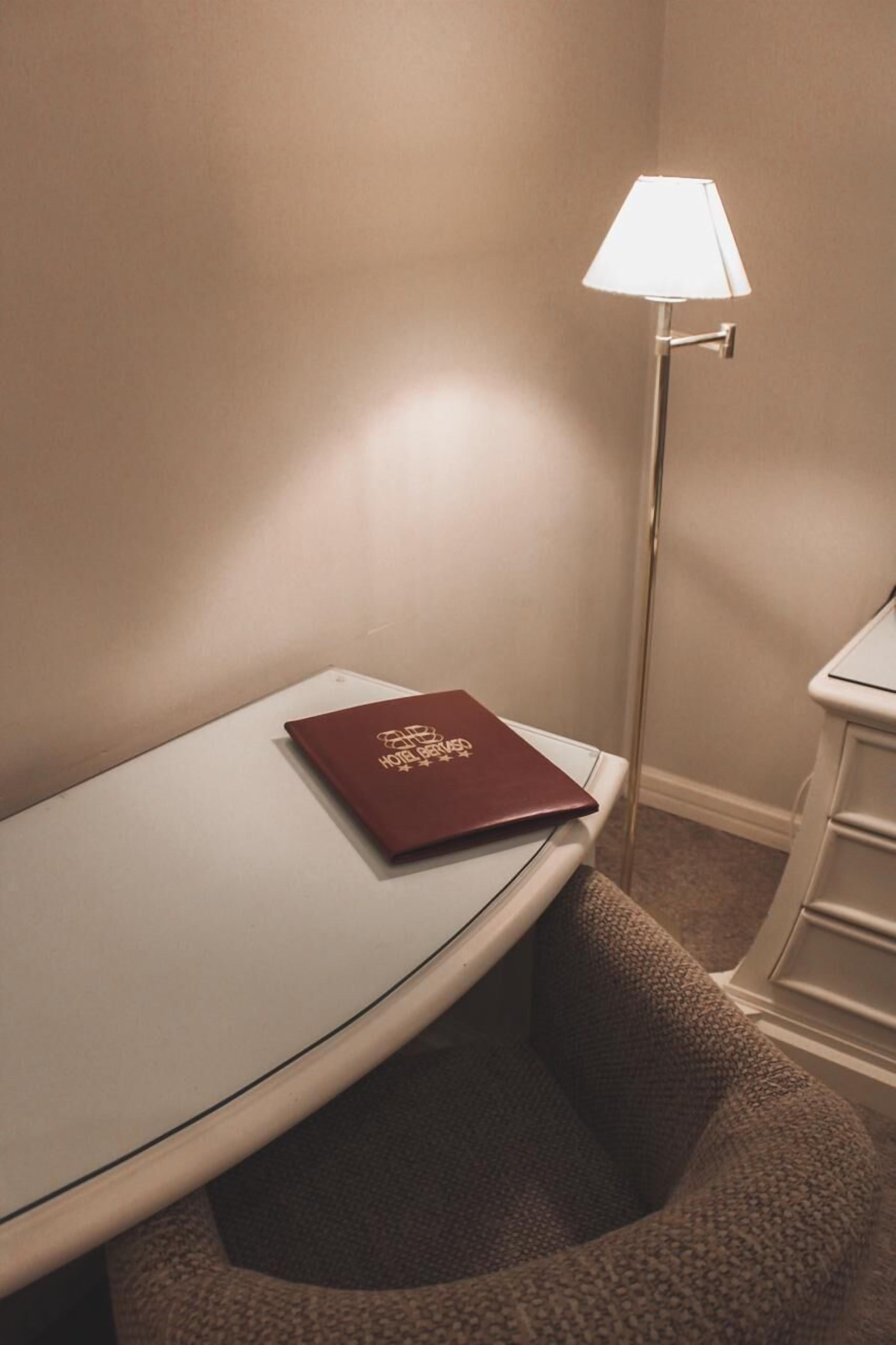 Een hoek van een hotelkamer met een klein tafeltje, een lamp en een rood boekje op het tafeltje.