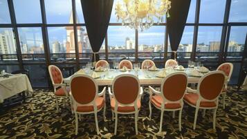 Comedor con una gran mesa dispuesta para seis personas, rodeada de sillas con cojines naranja, una araña y vistas a un paisaje urbano a través de amplios ventanales.