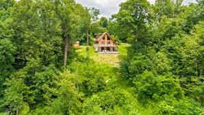Une cabane en bois entourée d’une forêt dense, avec une terrasse visible et un chemin menant à l’entrée.