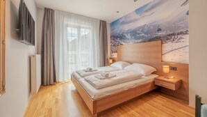 Een moderne hotelkamer met een houten bed, een groot raam met dunne gordijnen, een tv aan de muur en een wanddecoratie met een wintersportthema.