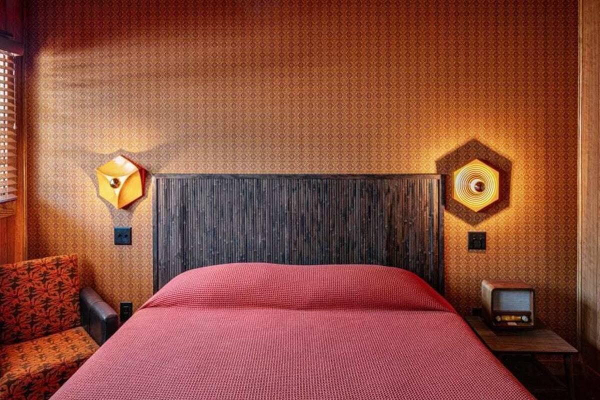 Una habitación de hotel con una cama grande, cabecera de madera, papel tapiz estampado y una mesita con una radio antigua.