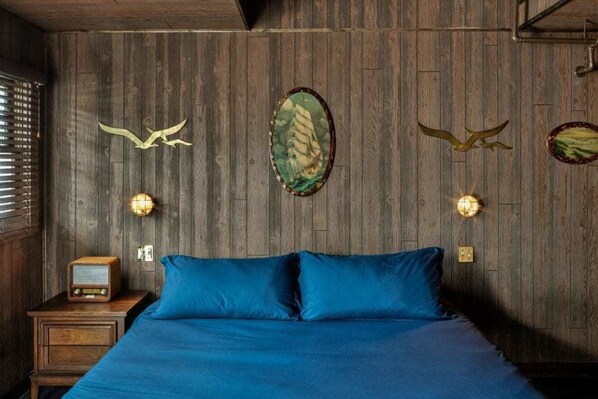 Un dormitorio con pared de madera, cama con ropa de cama azul, una radio antigua y luces fijadas en la pared.
