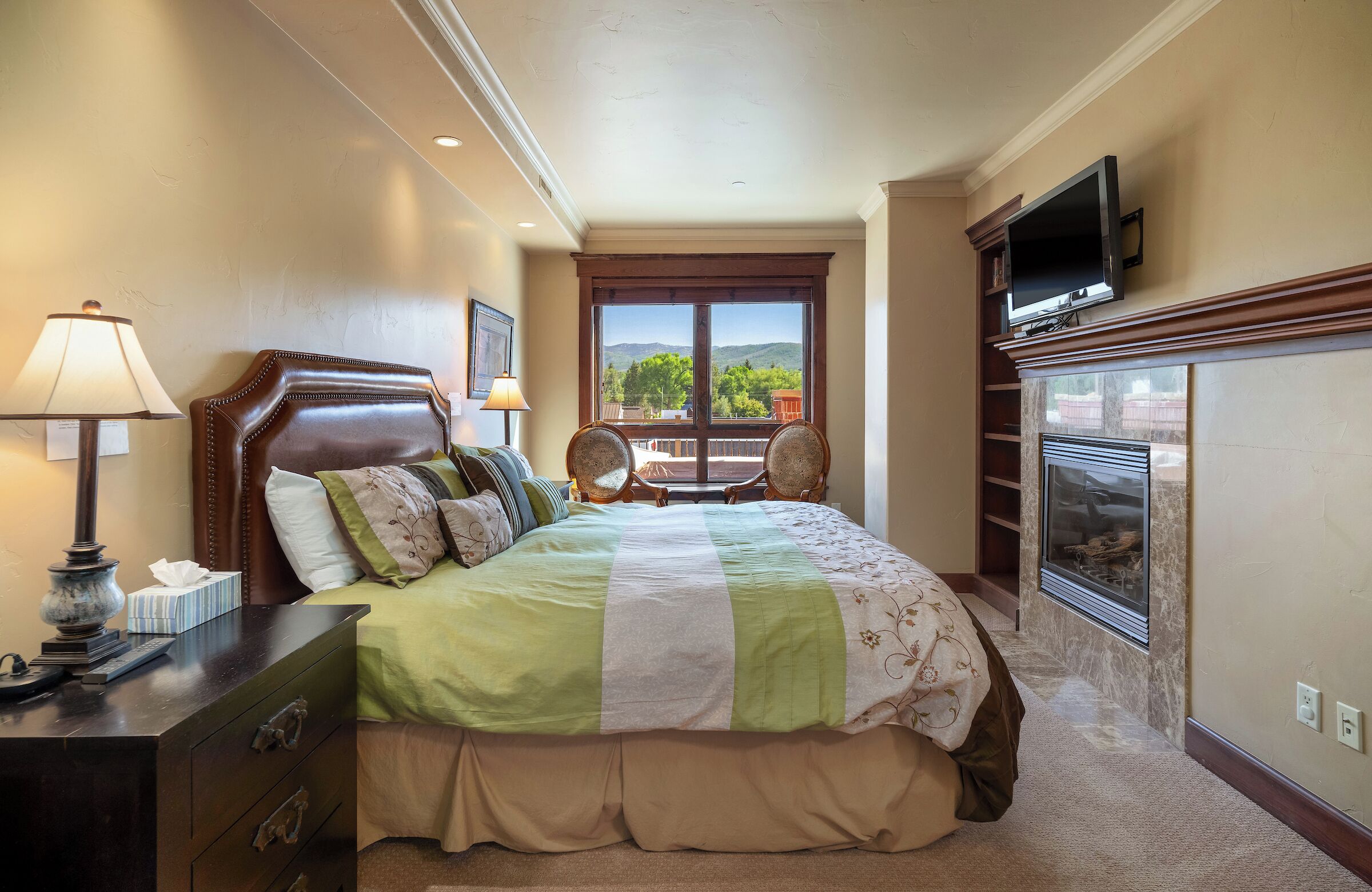 A hotel room with a large bed, a television mounted above it, a fireplace, and a view of the outdoors through a large window.
