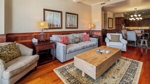 Ein geräumiges Wohnzimmer mit grauem Sofa, Holztisch und gemusterten Teppichen. Im Hintergrund ist eine Küche erkennbar.