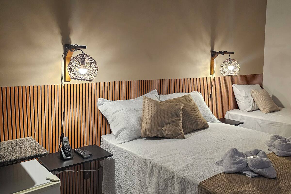 Quarto de hotel com duas camas, uma cabeceira de madeira, um minibar e luminárias fixadas na parede.