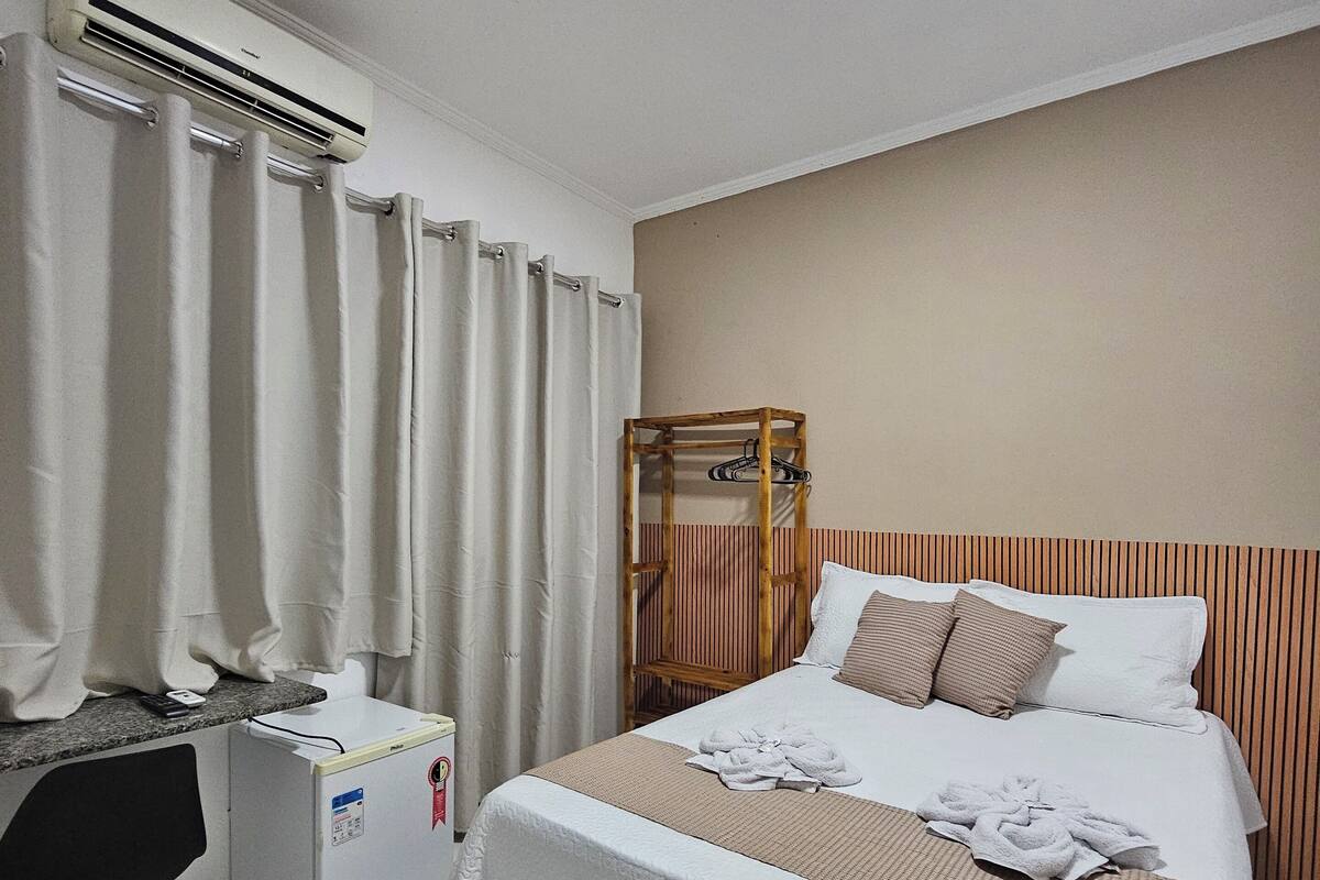 Quarto de hotel com uma cama, escrivaninha, ar condicionado e cabeceira de madeira.