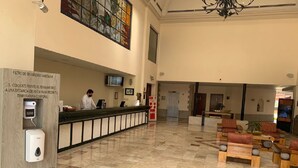 Un vestíbulo de hotel con mostrador de recepción, dispensador de gel hidroalcohólico, un amplio ventanal de vitral y una zona de estar con sillones acolchados.
