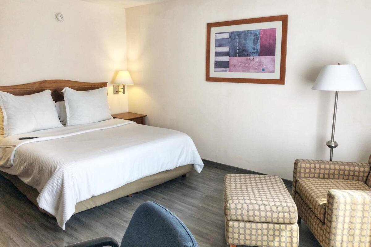 A hotel room with a bed, bedside table, lamp, chair, ottoman, and a framed abstract painting on the wall.