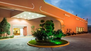 Una entrada de hotel con fachada curva, un jardín ajardinado con topiarios y una entrada bien iluminada.