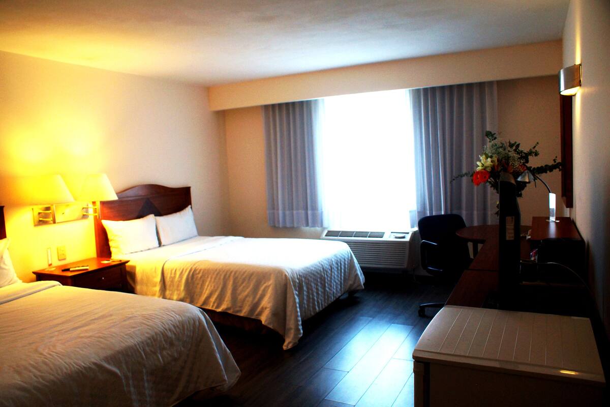 A hotel room with two beds, a desk, a chair, a lamp, and a window with curtains.