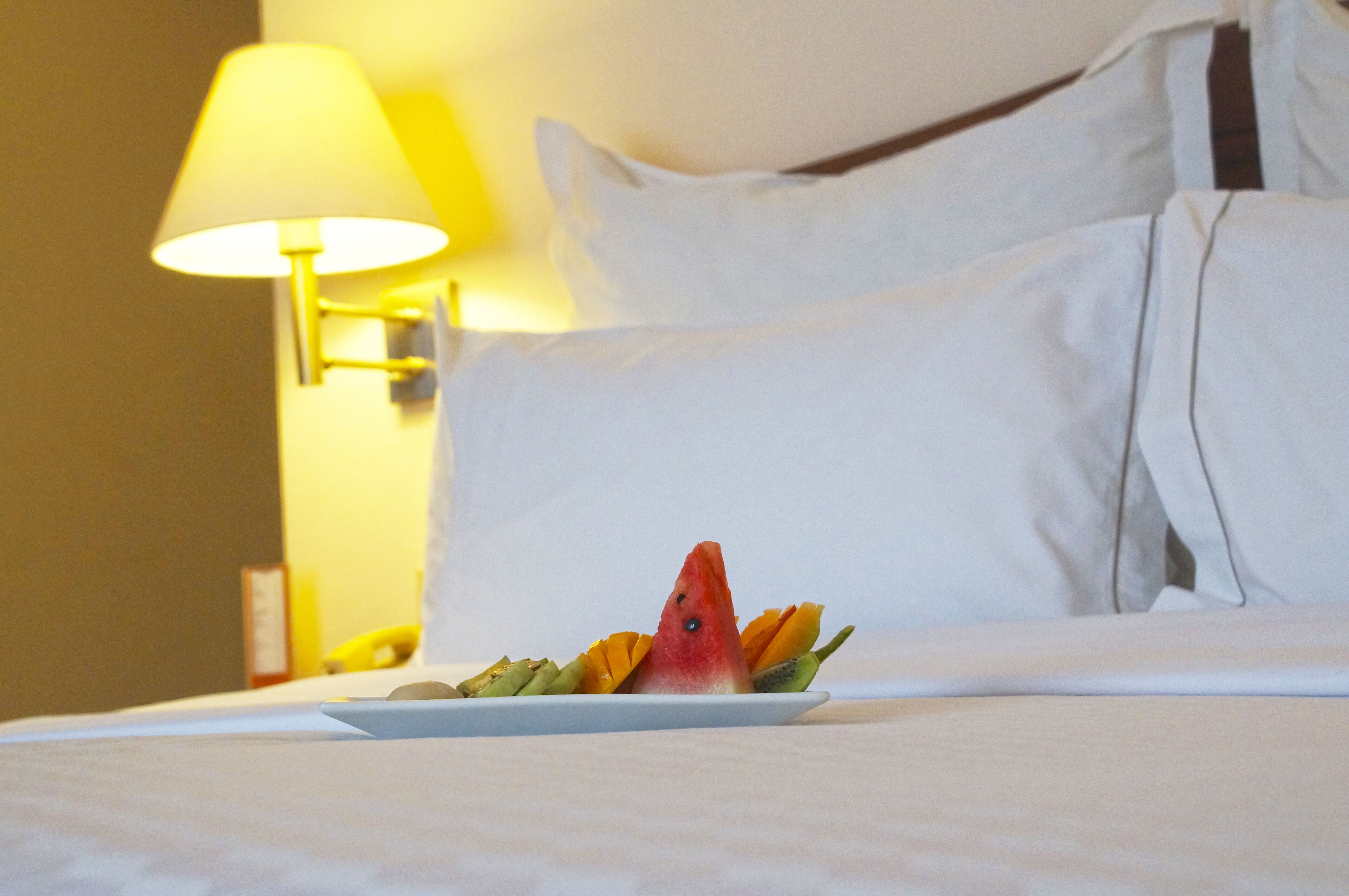 Una cama bien hecha con sábanas y almohadas blancas, una mesita de noche con una lámpara, y un plato de fruta que incluye una rebanada de sandía.