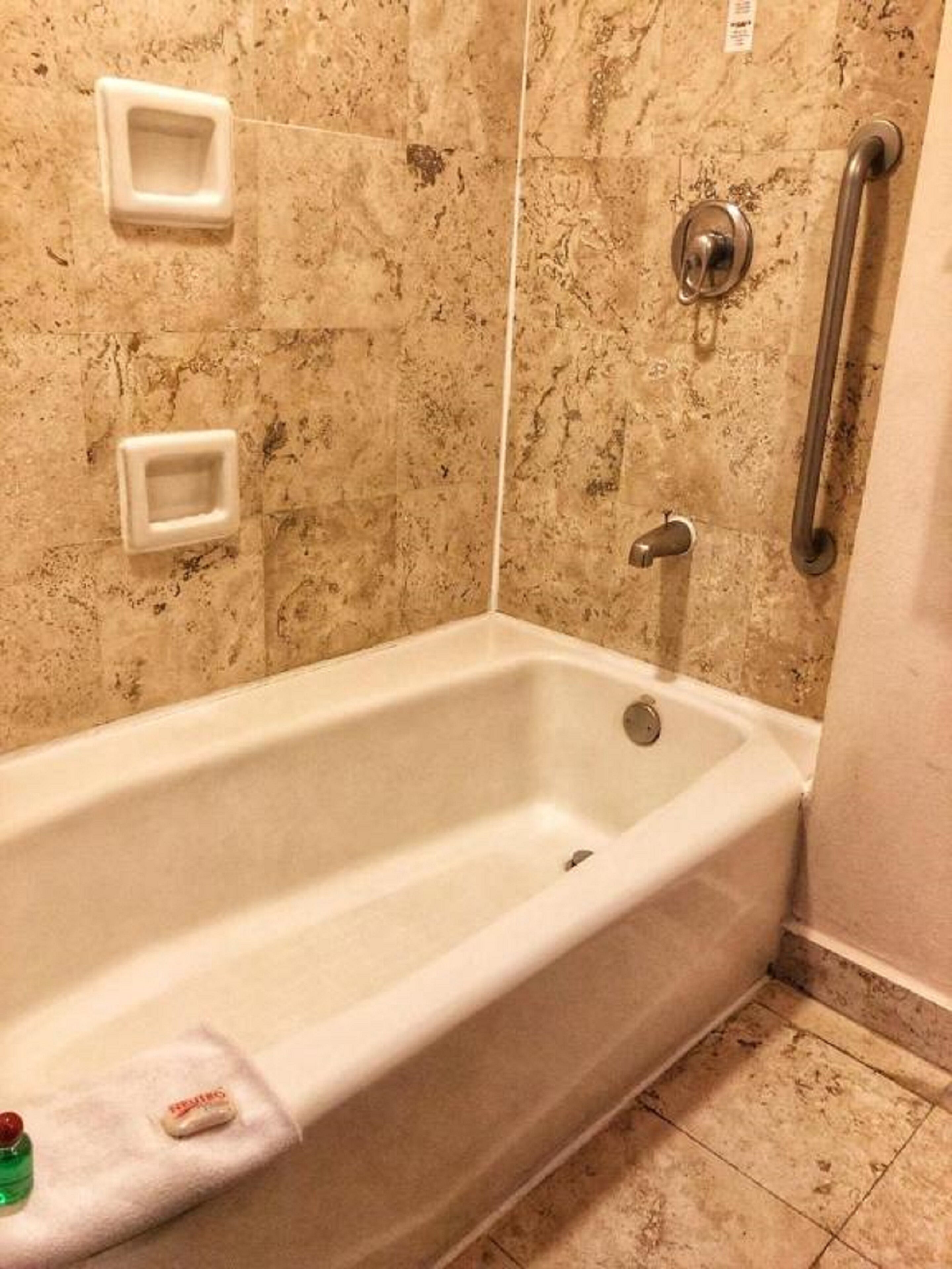 Una bañera con un cabezal de ducha manual, un asidero y dos dispensadores de jabón cuadrados en la pared.