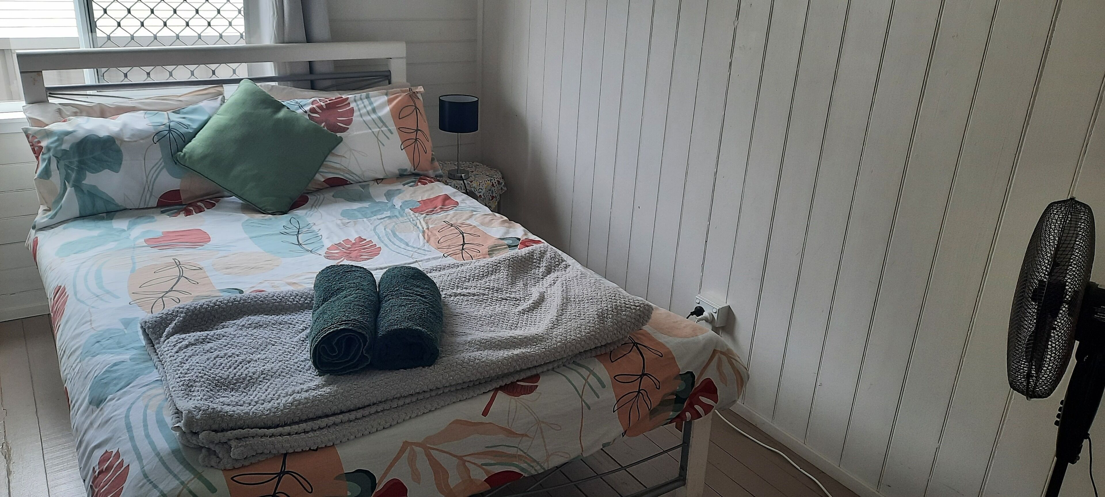 A neatly made bed with floral bedding, a green pillow, and two rolled towels.
