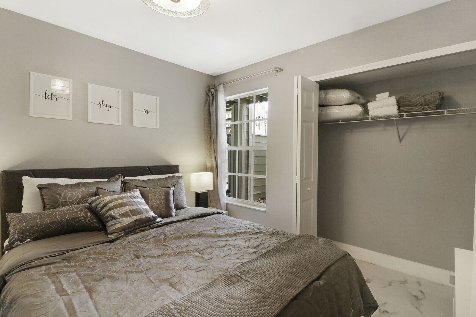 A bedroom with a bed, a bedside table, a nightstand, a window with curtains, a closet with shelves, and wall art.
