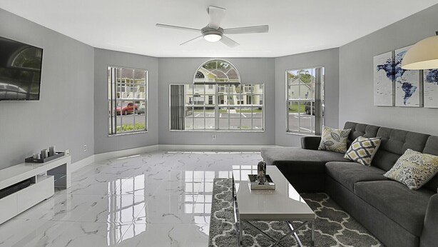 A modern living room with a grey couch, a coffee table, and a TV mounted on the wall. There are large windows providing a view of the street and houses outside. - Completely Renovated 3/2 Close to Disney parks (Kissimmee)