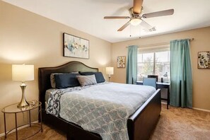 Un dormitorio con una cama, dos lámparas de noche, un ventilador de techo, una ventana con cortinas y obras de arte en las paredes.