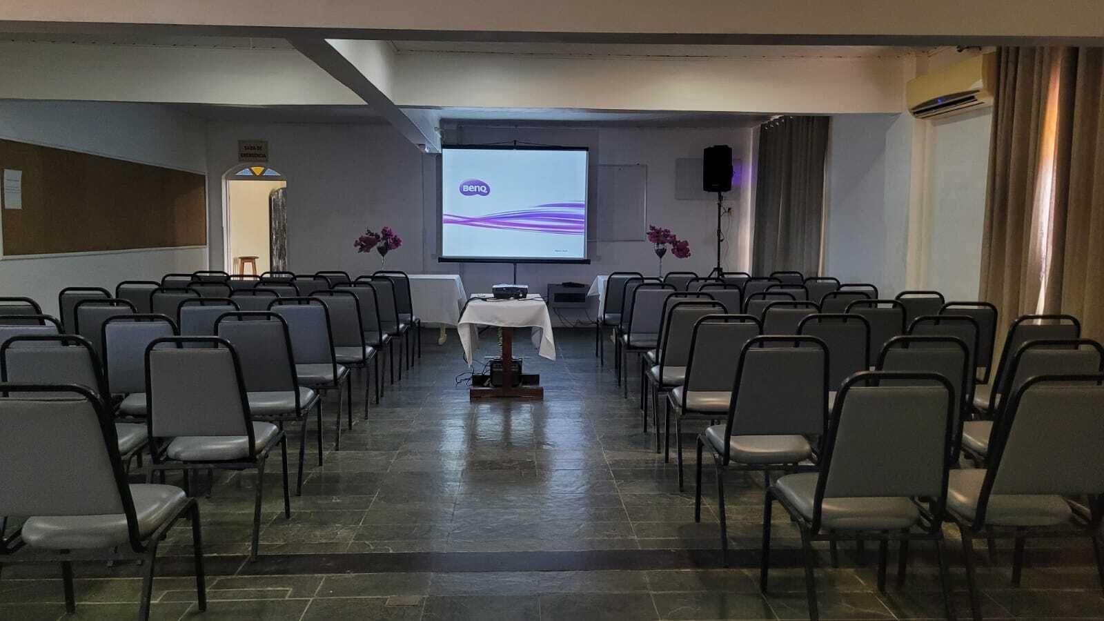 A conference room with rows of chairs, a podium, a screen, and a speaker.